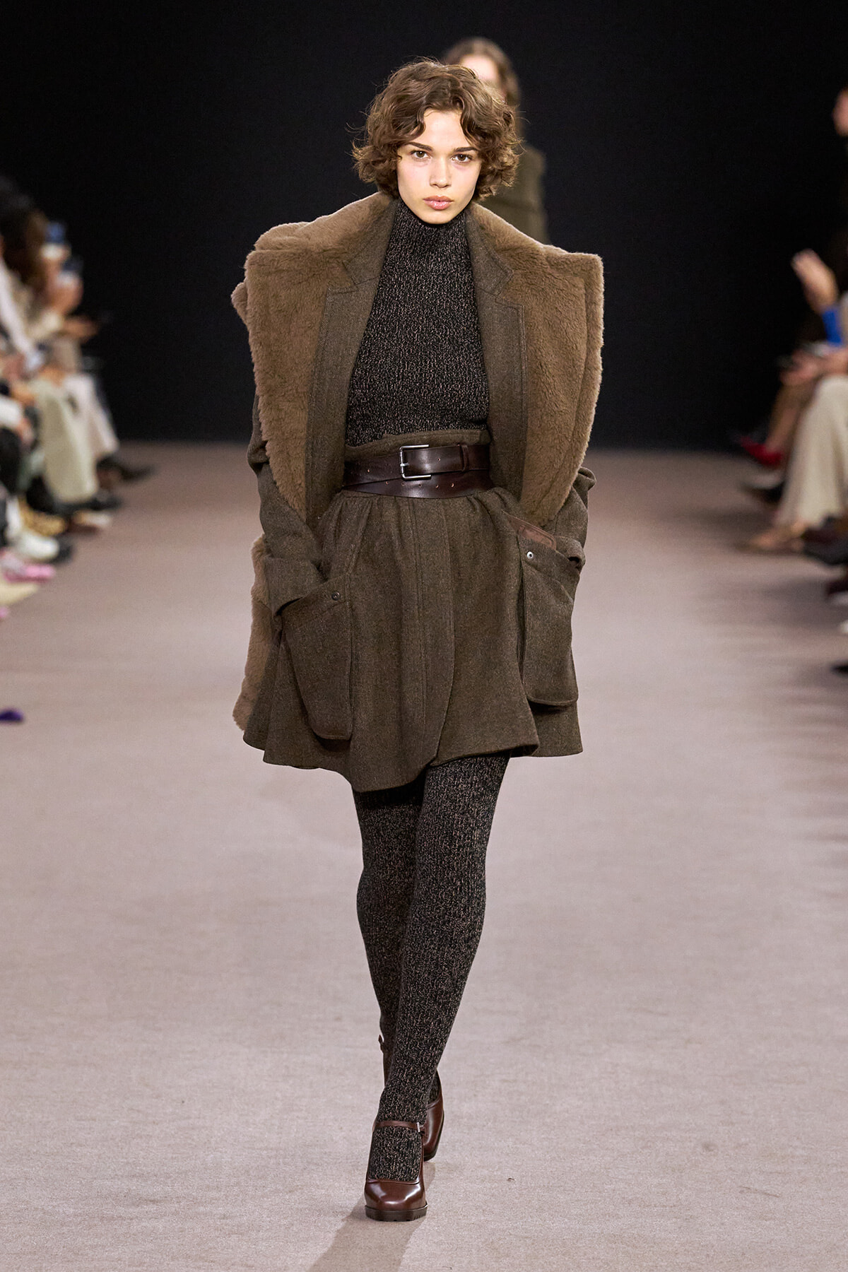 Model walking runway in an oversized brown shearling coat over a dark knit turtleneck, wide leather belt, and textured tights with brown heels.