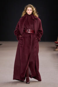 Model on a runway wearing a long burgundy faux fur coat with a wide belt and matching turtleneck.