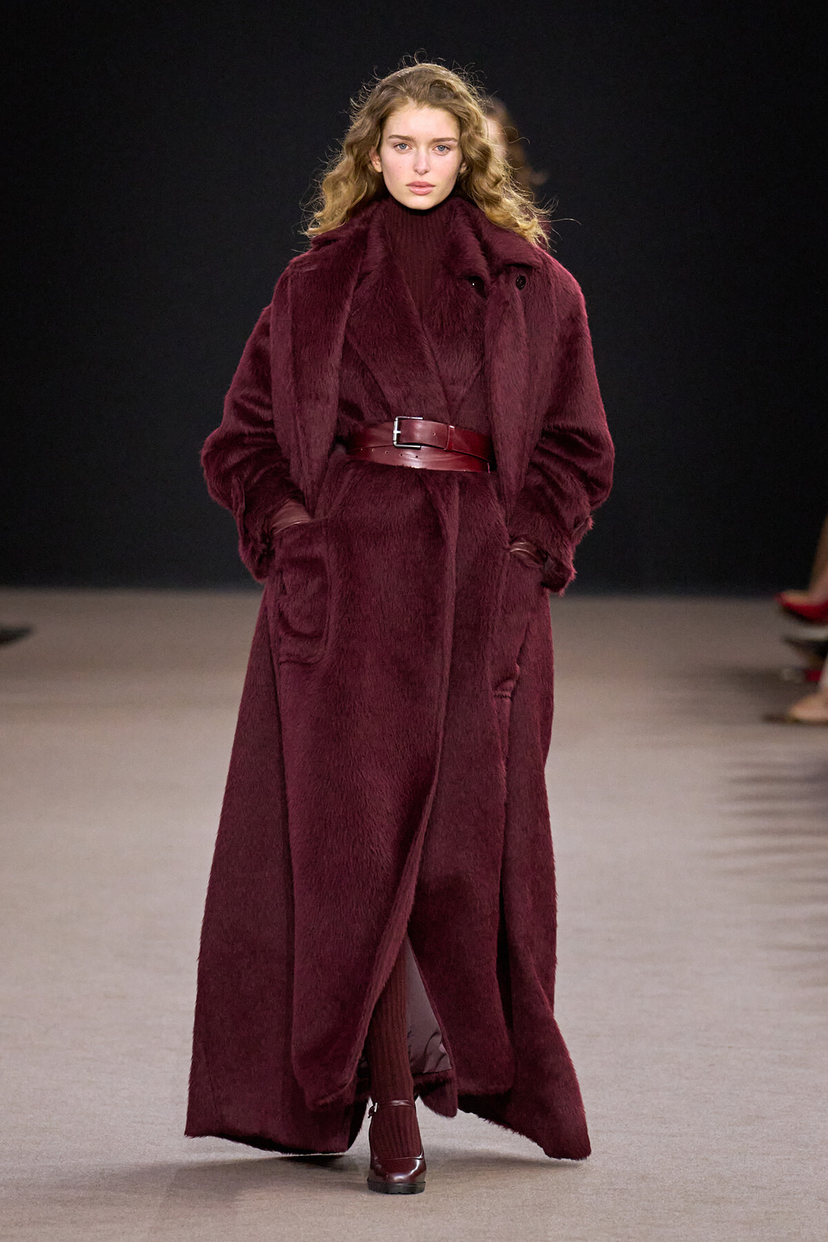 Model on a runway wearing a long burgundy faux fur coat with a wide belt and matching turtleneck.