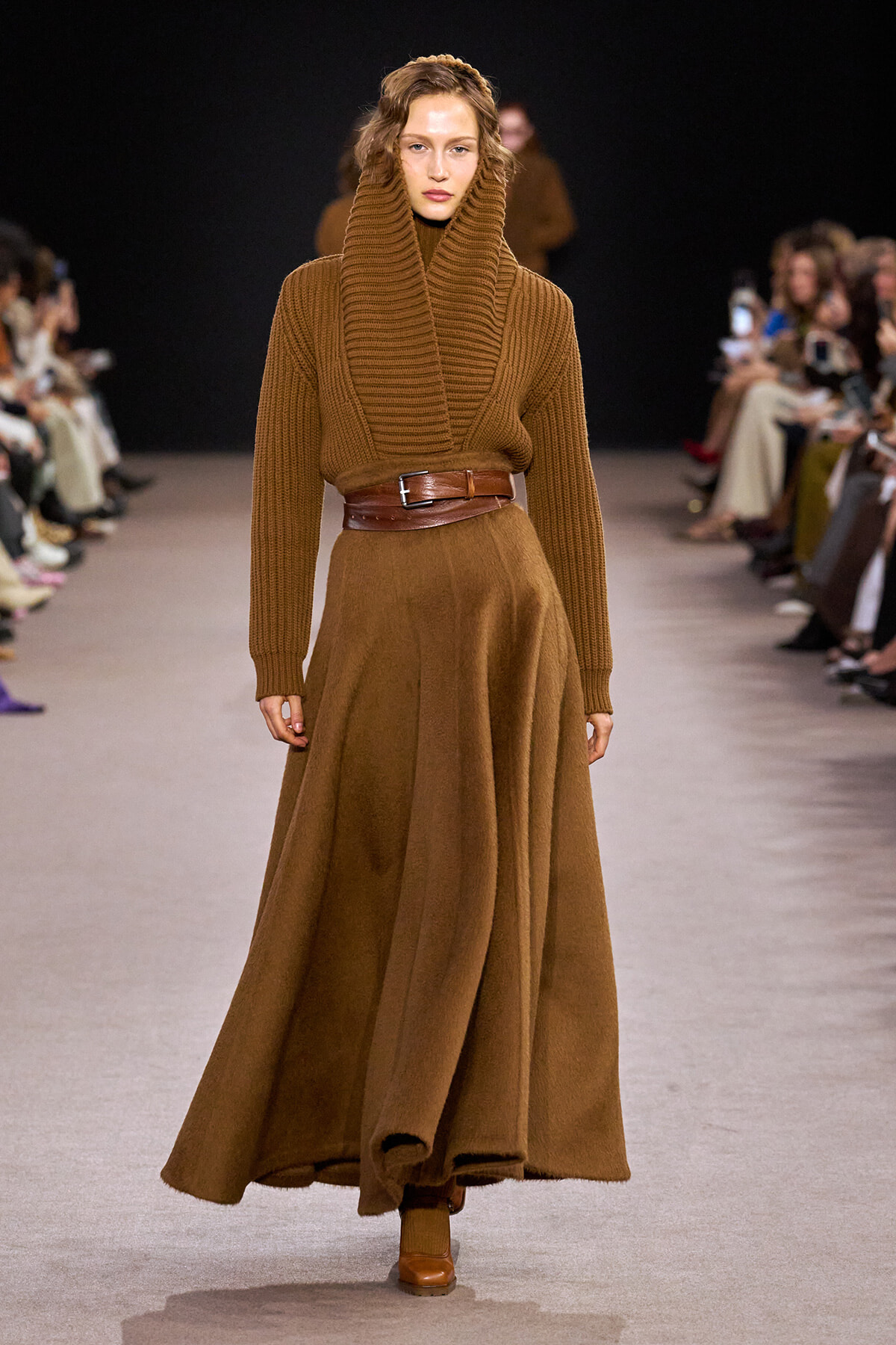 Model walks runway in a chunky brown turtleneck sweater, wide brown belt, and long camel-colored skirt with brown boots.