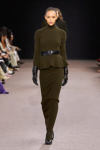 Model on a runway wearing an olive green ribbed knit turtleneck peplum top with a matching skirt and a wide black belt, plus black gloves and heels.