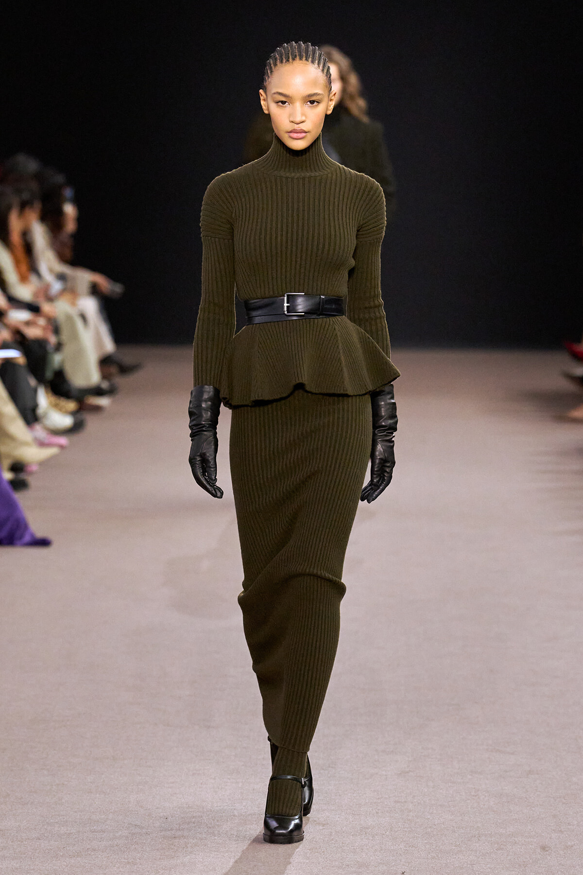 Model on a runway wearing an olive green ribbed knit turtleneck peplum top with a matching skirt and a wide black belt, plus black gloves and heels.