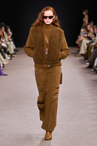 Model on a runway wearing a brown shearling vest over a brown turtleneck, with wide-leg brown pants and tan boots, accessorized by sunglasses.