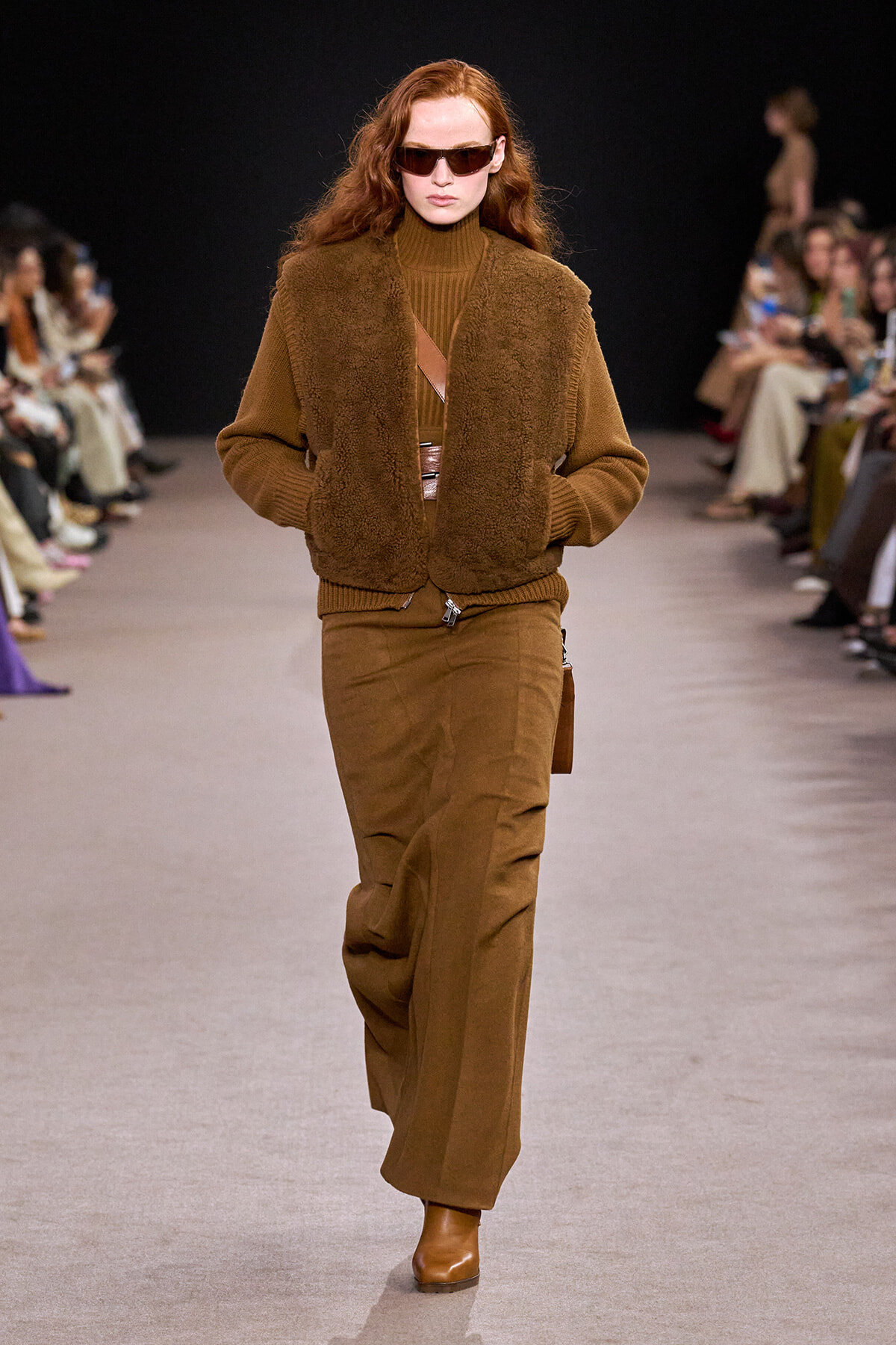 Model on a runway wearing a brown shearling vest over a brown turtleneck, with wide-leg brown pants and tan boots, accessorized by sunglasses.