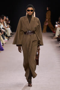 Model walks the runway in a brown oversized cape coat belted at the waist with dark sunglasses.