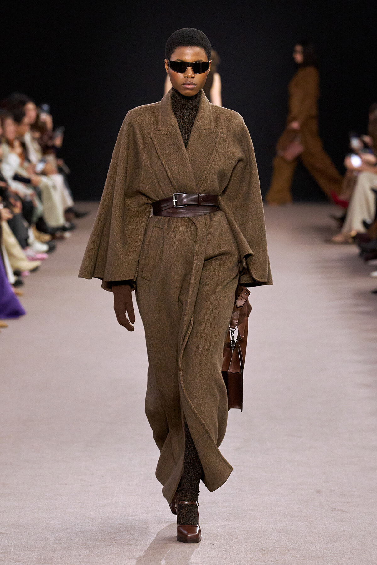 Model walks the runway in a brown oversized cape coat belted at the waist with dark sunglasses.