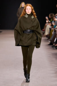 Model walking a fashion runway in an oversized olive-green cape coat cinched with a wide black belt, paired with ribbed olive tights and black ankle boots.