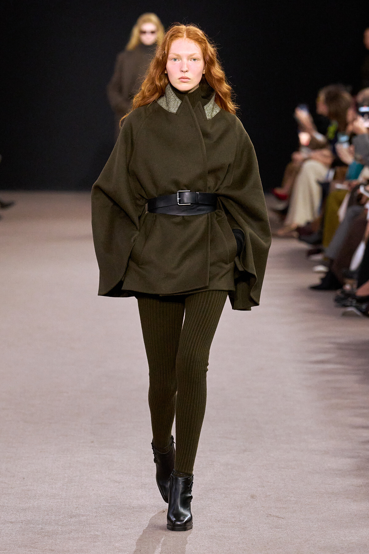 Model walking a fashion runway in an oversized olive-green cape coat cinched with a wide black belt, paired with ribbed olive tights and black ankle boots.