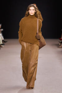 Model walking the runway in a chunky camel-brown turtleneck sweater and wide tan leather pants, carrying a brown shoulder bag.