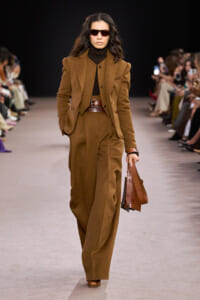 Female model walks a brown tailored suit ensemble with wide-shoulder blazer, matching vest, and wide brown belt, carrying a leather brown bag; wearing dark sunglasses on a runway.