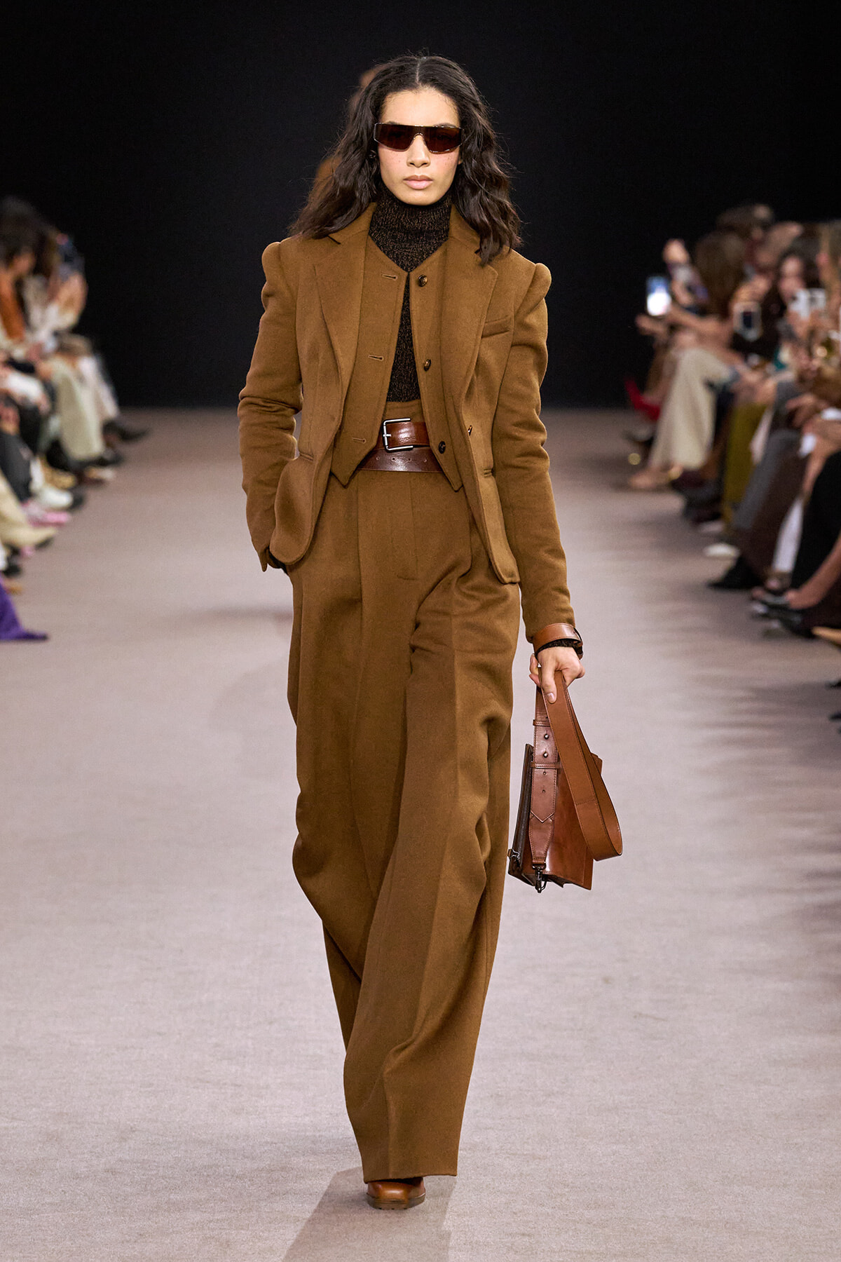 Female model walks a brown tailored suit ensemble with wide-shoulder blazer, matching vest, and wide brown belt, carrying a leather brown bag; wearing dark sunglasses on a runway.