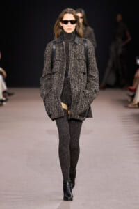 Model walking the runway in a chunky textured gray-black coat with large pockets, belt, black tights, and ankle boots, wearing dark sunglasses.