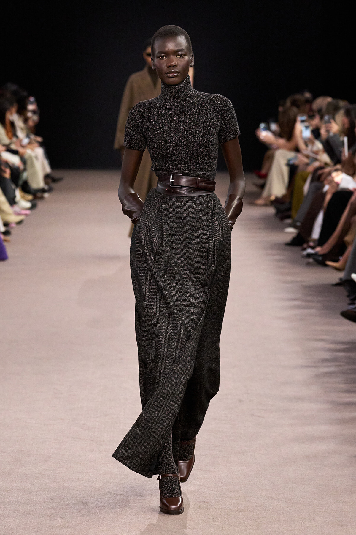 Model walks a fashion runway wearing a dark, textured turtleneck top with matching wide-leg pants cinched by a wide brown belt and brown gloves. Audience lines the catwalk in the background.