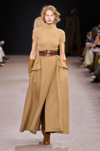 Model walking the runway in a tan ribbed turtleneck and a voluminous camel wrap skirt with a wide brown belt and brown gloves.