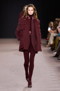 Model walking a burgundy monochrome outfit on a runway, wearing a oversized coat over a turtleneck and ribbed pants with matching burgundy accessories