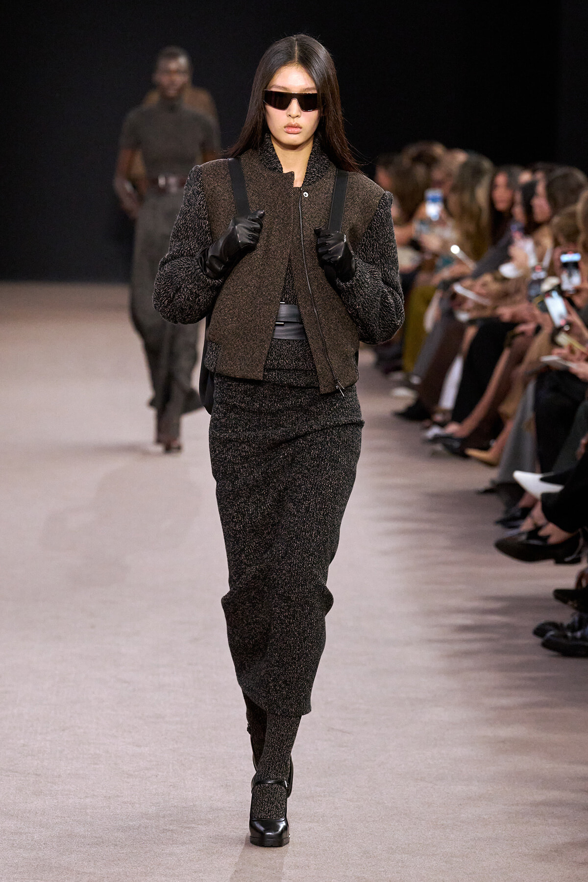 Model walking a fashion show runway in a dark textured two-piece suit, black gloves, and futuristic sunglasses.