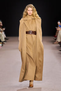 Model walking the runway in a camel oversized cape-style coat cinched with a wide brown belt; monochrome tan ensemble with matching turtleneck and boots.