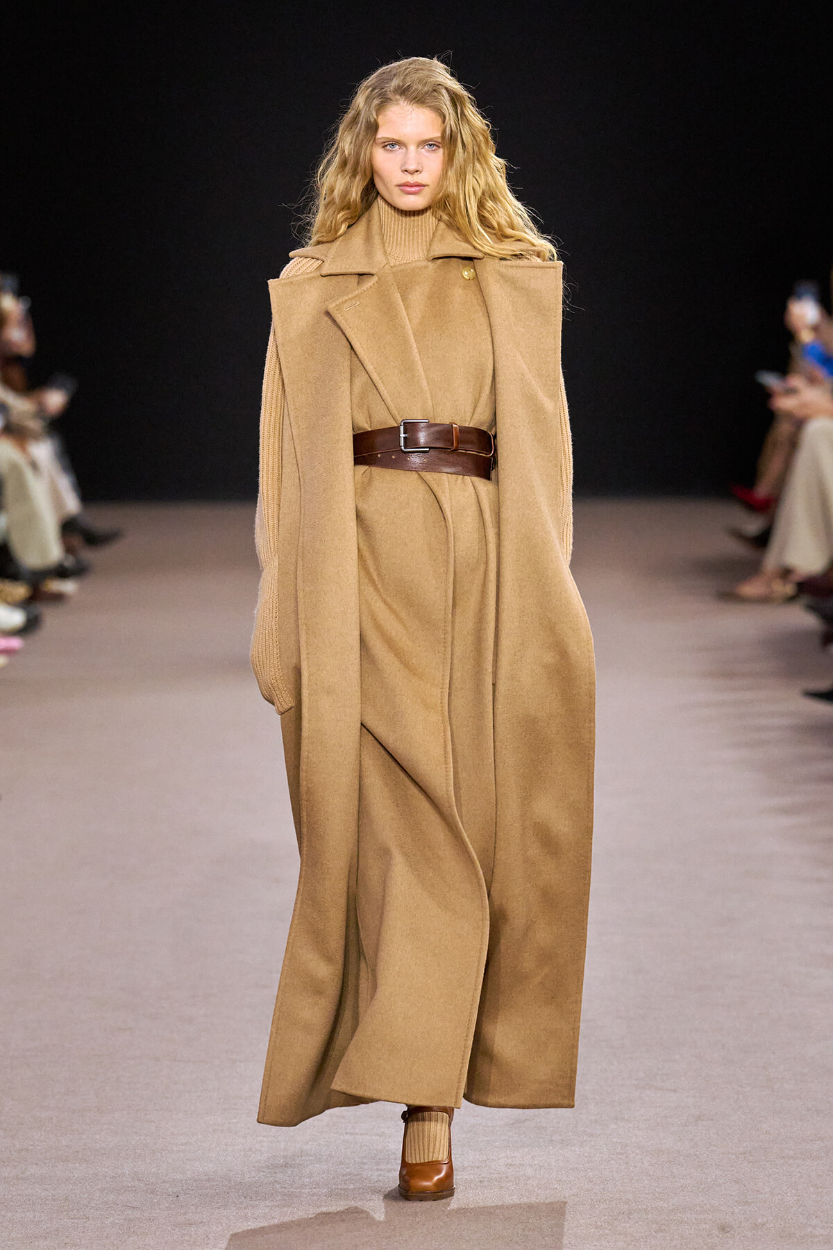 Model walking the runway in a camel oversized cape-style coat cinched with a wide brown belt; monochrome tan ensemble with matching turtleneck and boots.