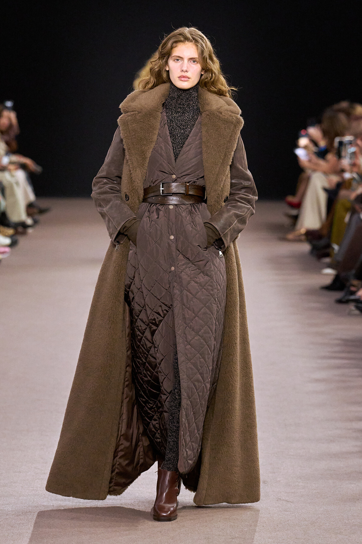 Model walking a runway in a brown shearling coat with a quilted inner layer, belted at the waist, and brown ankle boots.