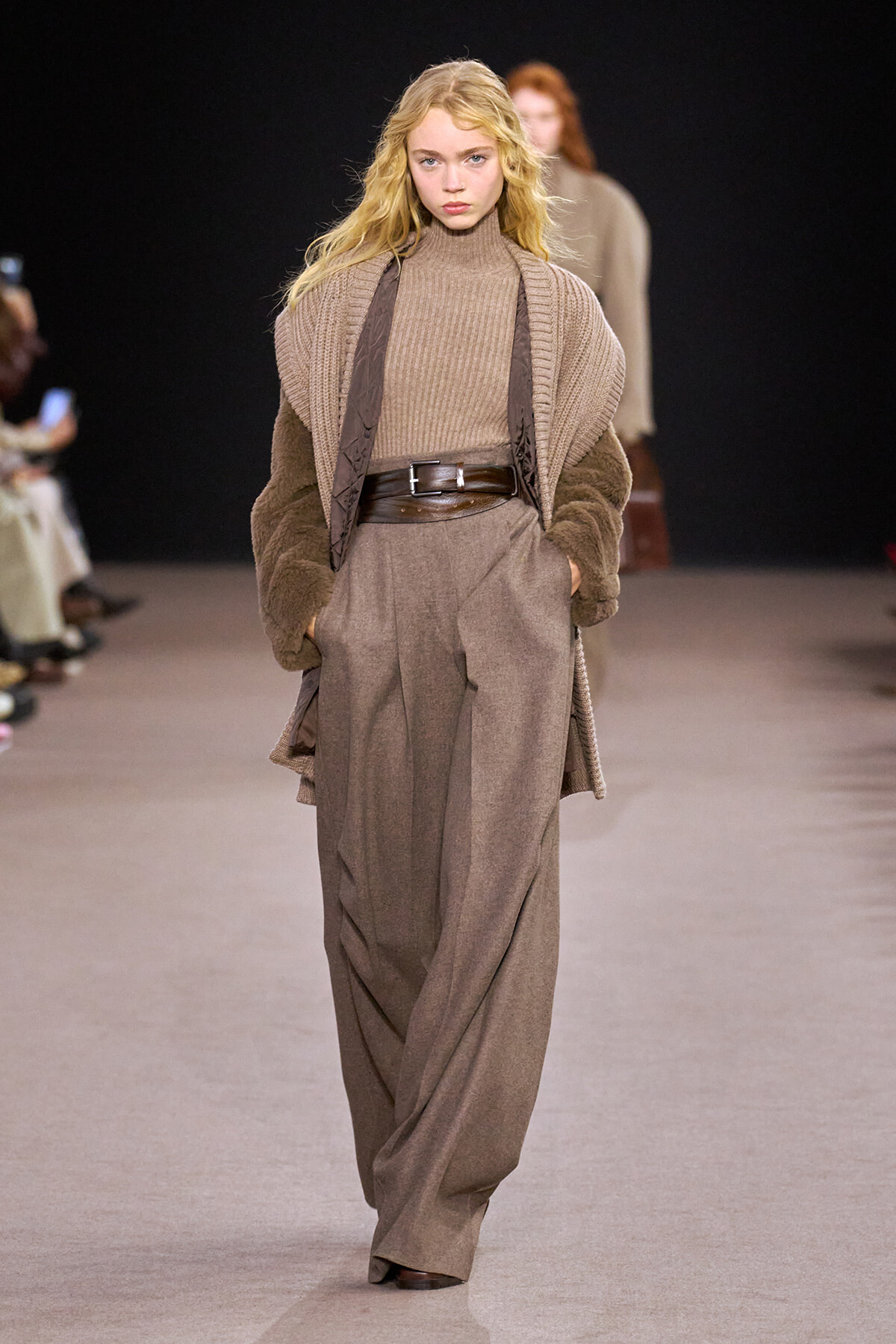 Model walks the runway in a head-to-toe beige outfit: chunky knit turtleneck, oversized cardigan with fur sleeves, a wide brown belt, and wide-leg taupe trousers.