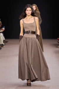 Model walks the runway in a sleeveless taupe knit top with a wide dark brown belt and a full, flowing taupe skirt, paired with brown gloves.