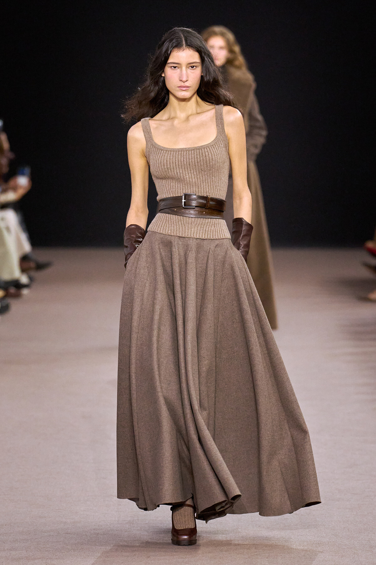 Model walks the runway in a sleeveless taupe knit top with a wide dark brown belt and a full, flowing taupe skirt, paired with brown gloves.