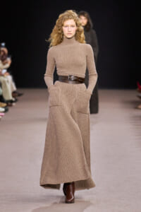 Model on a runway wearing a beige ribbed knit turtleneck dress, a wide brown belt, and brown boots, hands in pockets.