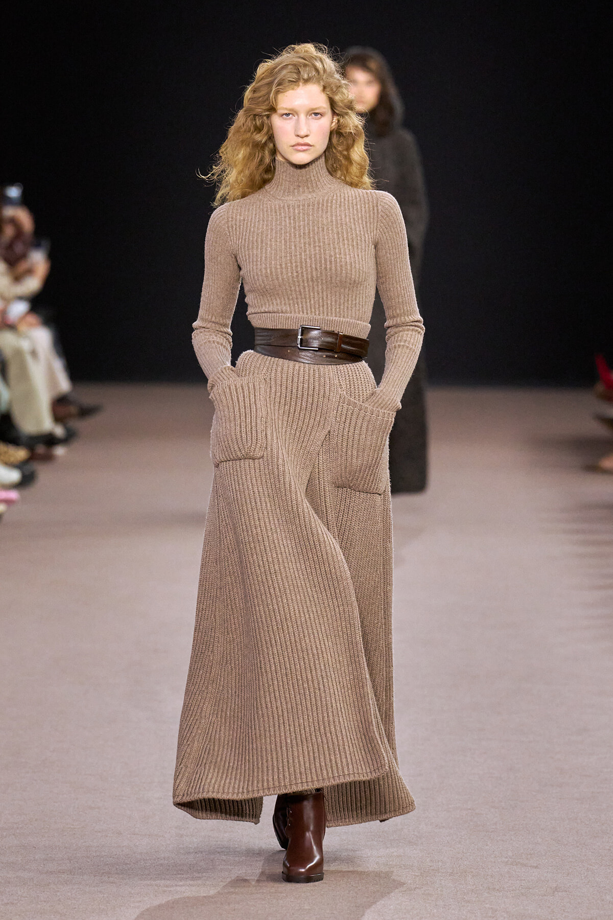 Model on a runway wearing a beige ribbed knit turtleneck dress, a wide brown belt, and brown boots, hands in pockets.