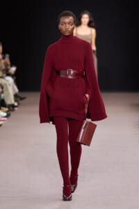 Model on a runway wearing a burgundy ribbed knit turtleneck dress with a wide belt and cape-like sleeves, carrying a brown rectangular handbag.