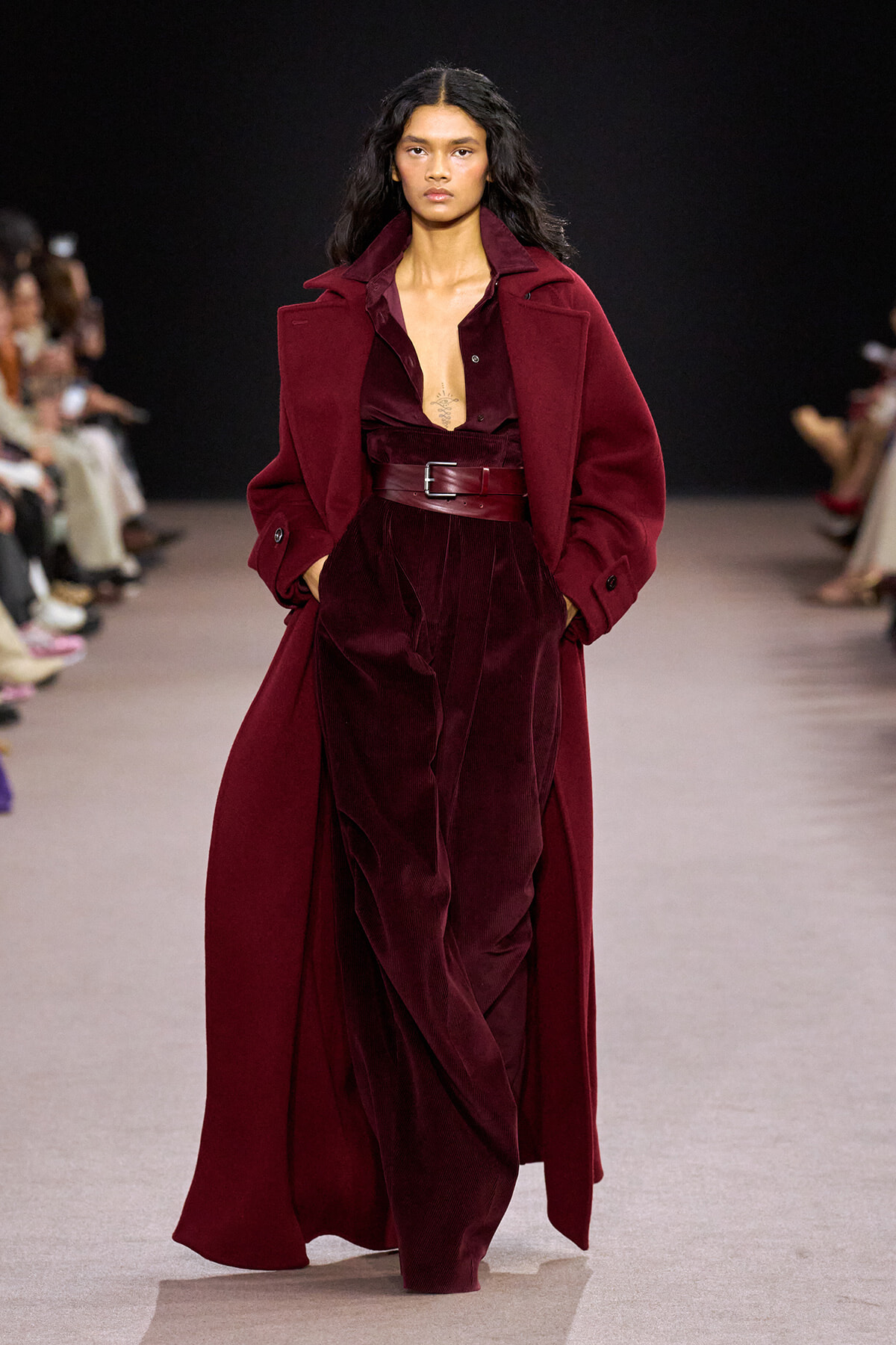 Model walks a runway wearing a burgundy velvet suit with a long matching coat and wide belt; black backdrop and seated audience on each side.