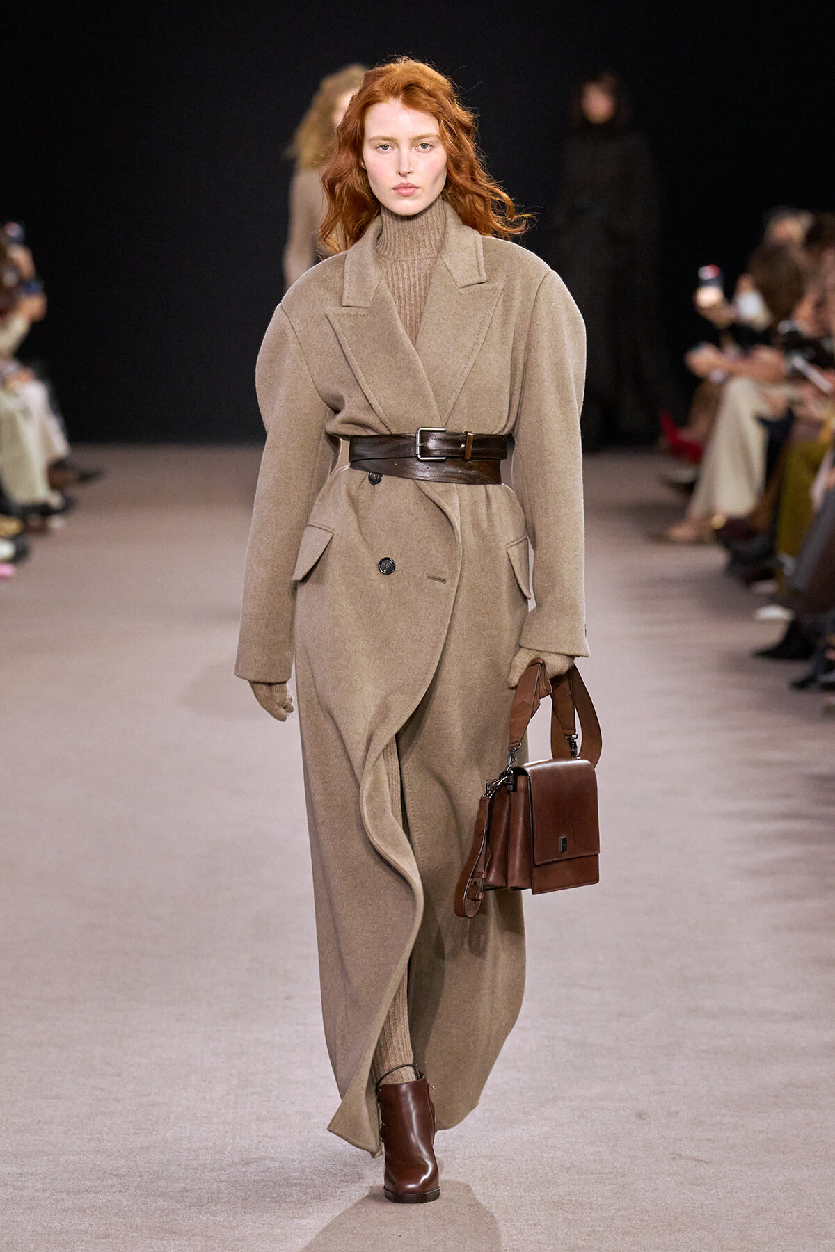 Model walks runway in a long beige wool coat belted at the waist, with a tan turtleneck and brown leather handbag and boots.