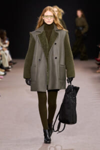 Model walks the runway in a green shearling-lined coat, turtleneck dress, gloves, and sunglasses, carrying a black backpack-like bag.