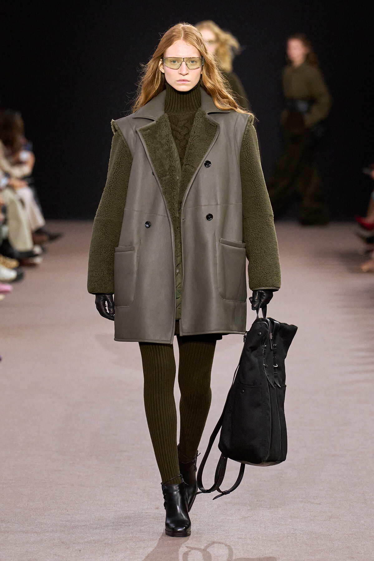 Model walks the runway in a green shearling-lined coat, turtleneck dress, gloves, and sunglasses, carrying a black backpack-like bag.