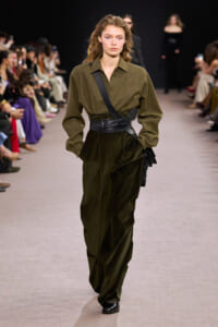 Model walks the runway in an olive-green coordinated outfit: button-up top, wide-leg pants, and a black wide belt with a diagonal shoulder strap and bag.