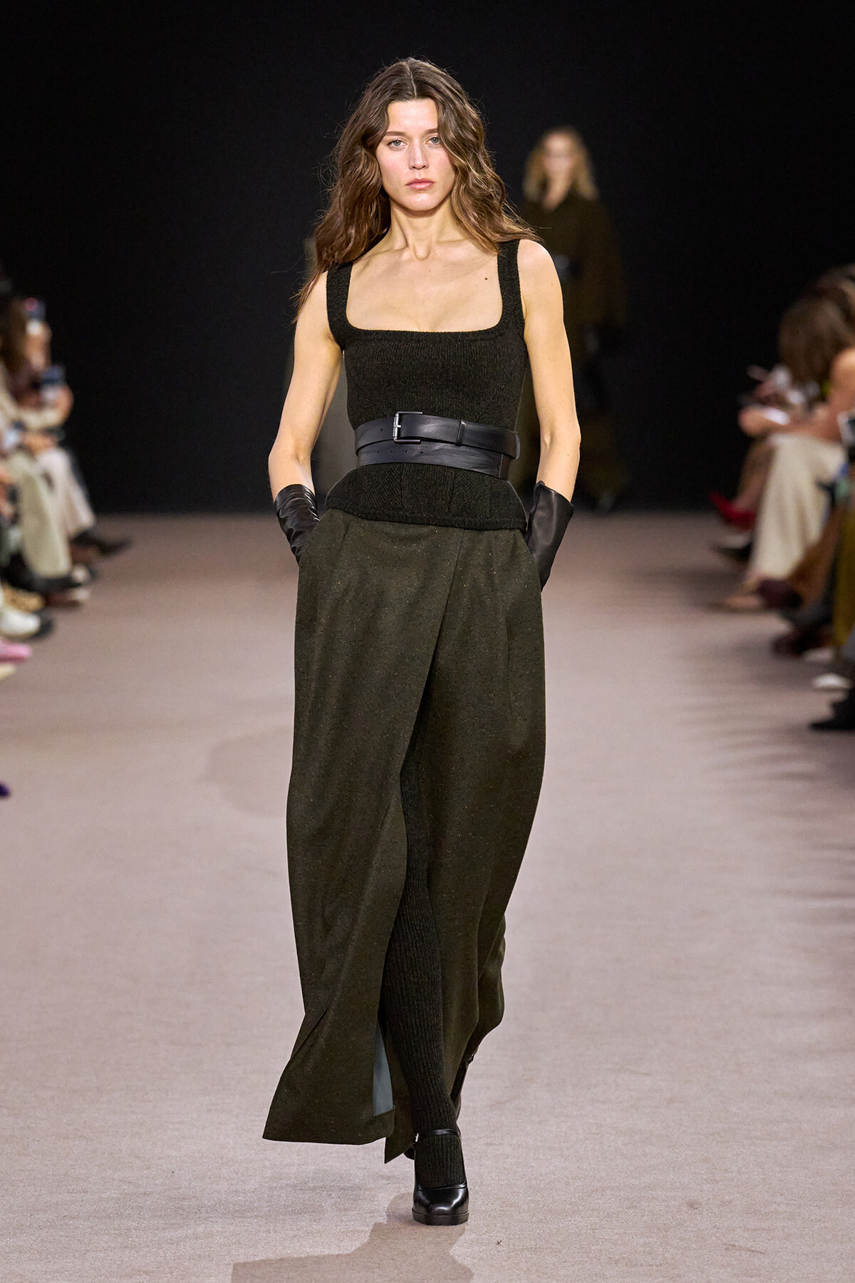 Model walking down runway in a dark, sleeveless square-neck top with a wide black belt over olive wide-leg pants and black gloves.