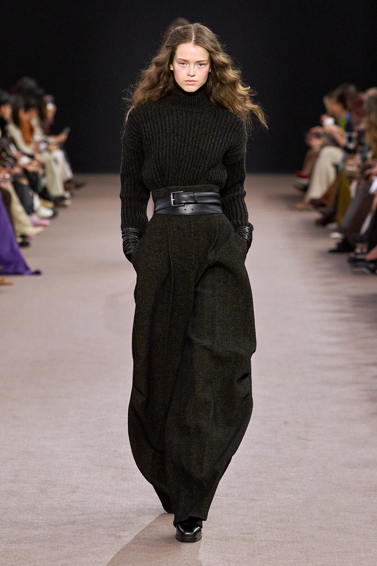 Model walks a runway in a black knit turtleneck with a wide belt and a long textured black skirt, wearing gloves.
