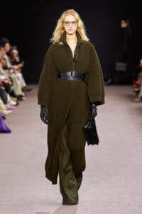 Model on a runway wearing an oversized olive-green knit coat belted at the waist with black gloves and a black clutch, sunglasses on.