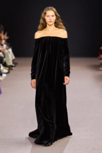 Model walking a long, off-the-shoulder black velvet gown on a fashion runway with a dark background and seated audience in the foreground.