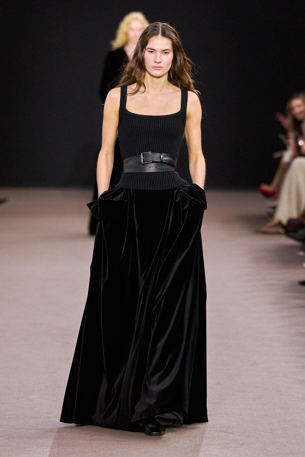 Model walking the runway in a black sleeveless ribbed top, wide belt, and a floor-length velvet skirt with hands in pockets.