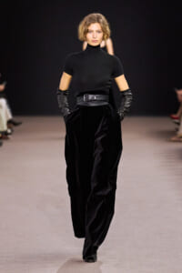 Model on a runway wearing an all-black outfit: black turtleneck top, wide velvet pants with a belt, and long black leather gloves, walking forward.