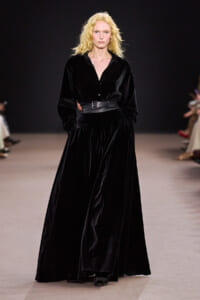 Model walks the runway in a floor-length black velvet dress with a wide waist belt and hands in pockets.