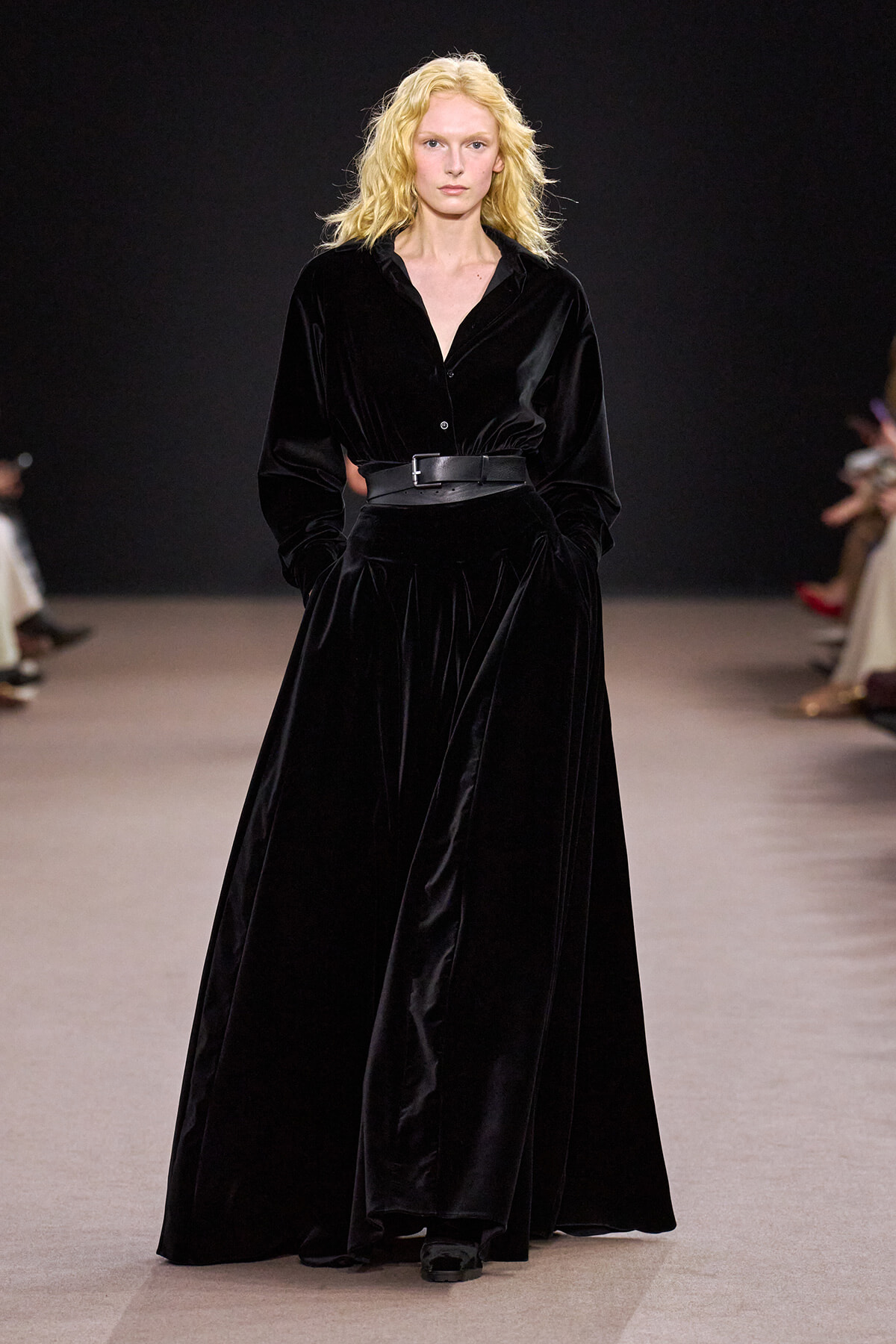 Model walks the runway in a floor-length black velvet dress with a wide waist belt and hands in pockets.