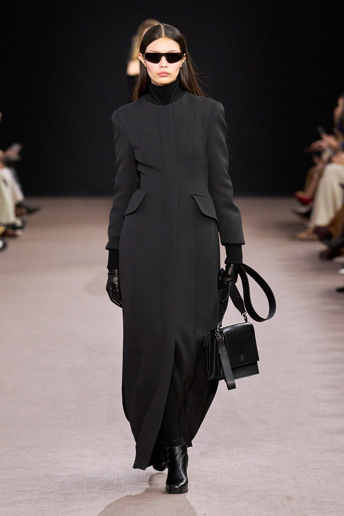 Model walking runway in a long black coat, black gloves, and dark sunglasses, carrying a black structured handbag.