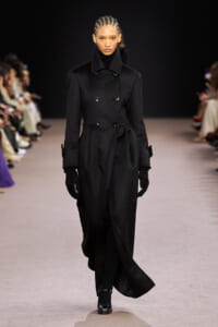 Model walking the runway in a long black double-breasted coat with gloves.