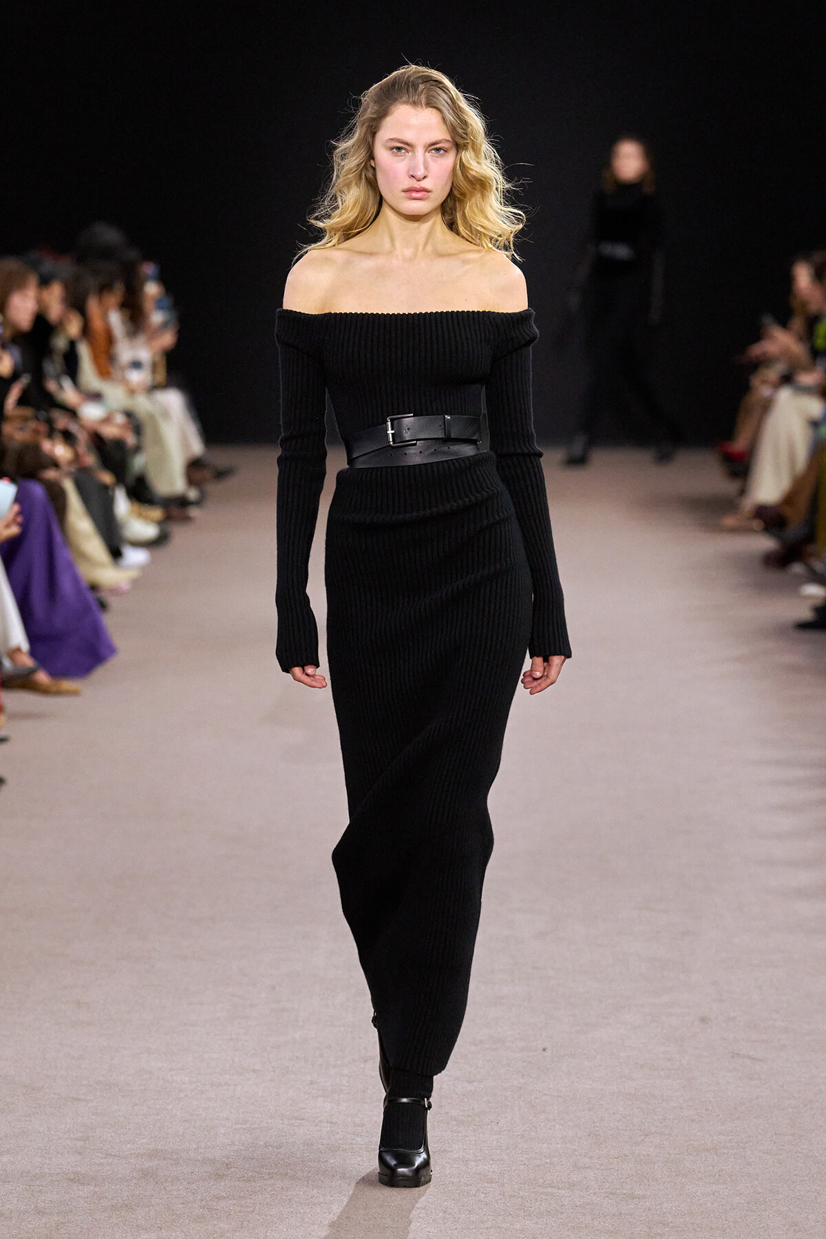 Model on a runway wearing an off-the-shoulder black ribbed knit dress with a wide black belt and ankle boots, audience blurred in the background.