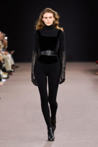 Model walks the runway in a black ribbed turtleneck jumpsuit with a wide belt and elbow‑length leather gloves.