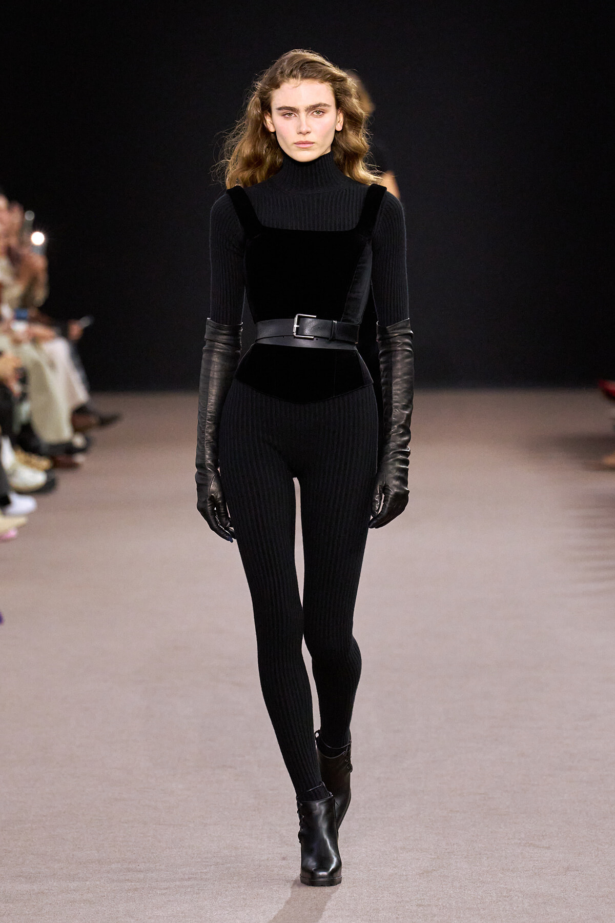 Model walks the runway in a black ribbed turtleneck jumpsuit with a wide belt and elbow‑length leather gloves.