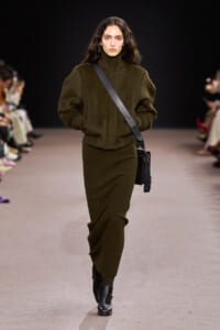 Model walking a runway in an olive green oversized knit sweater and matching ribbed skirt, with black ankle boots and a black crossbody bag.