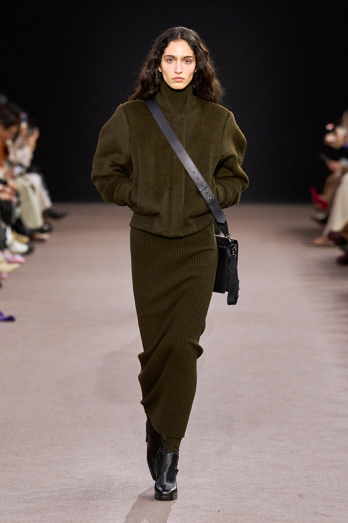 Model walking a runway in an olive green oversized knit sweater and matching ribbed skirt, with black ankle boots and a black crossbody bag.