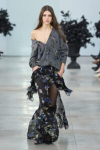Model on a fashion runway wearing an off-shoulder gray cardigan with floral embroidery and a sheer black floral-printed skirt.
